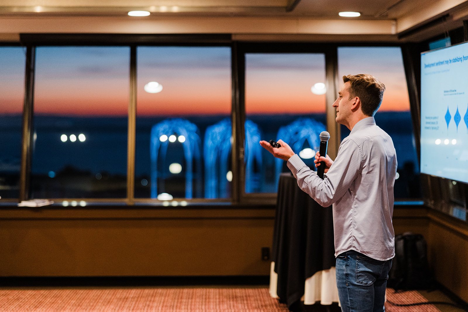 Brian Houck speaking with a handheld microphone in front of windows at sunset.